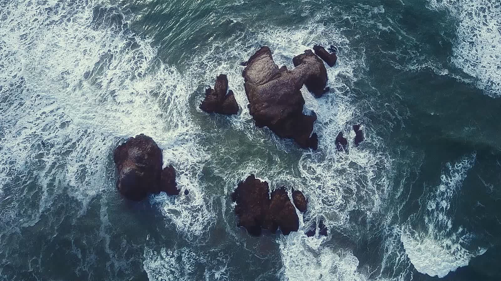Dark rocks seen from above, surrounded by churning white surf